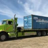 Metro FB-20 long-deck flatbed truck carrying a shipping container on job site