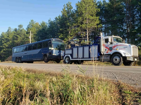 Metro Tow Trucks INT-16 white heavy-duty integrated wrecker towing passenger coach along roadside with trees in background