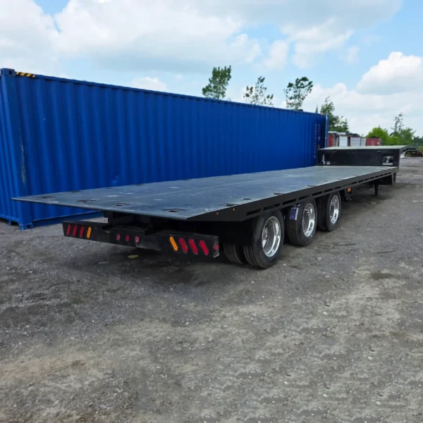 Side profile of STT-50 flatbed heavy-haul trailer with tri-axle setup parked beside shipping containers